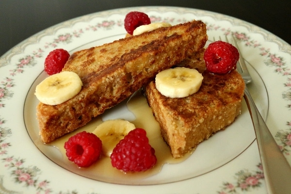 two pieces of french toast on a plate with syrup, sliced bananas, and raspberries