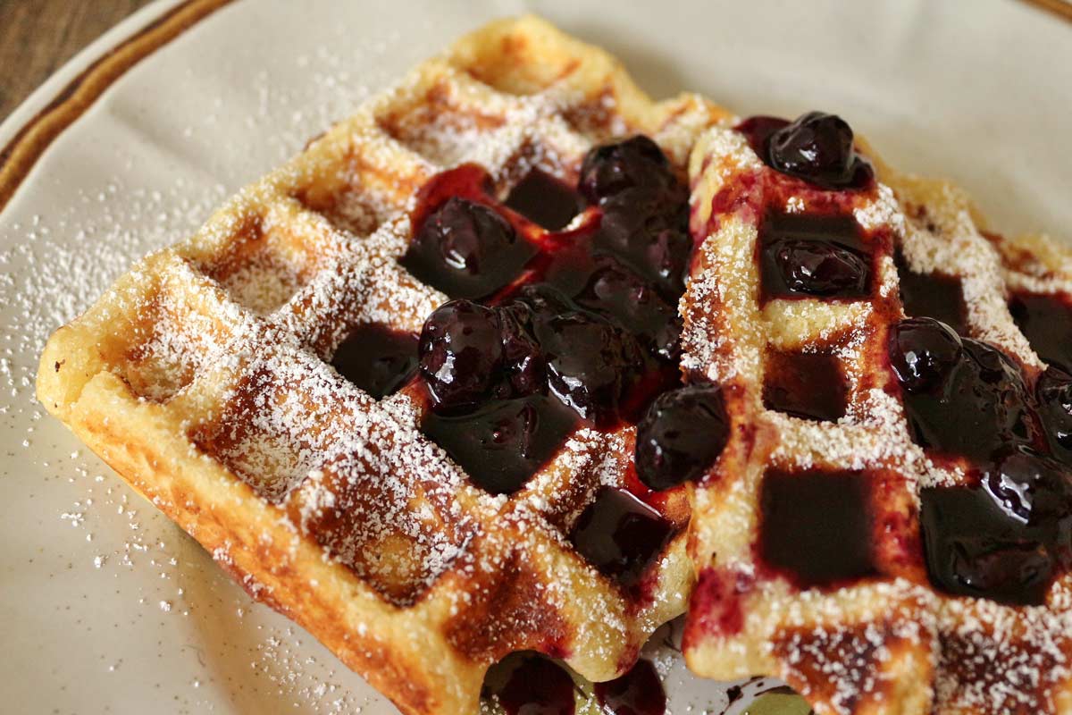 Two rectangular ricotta waffles topped with powdered sugar and blueberry syrup.