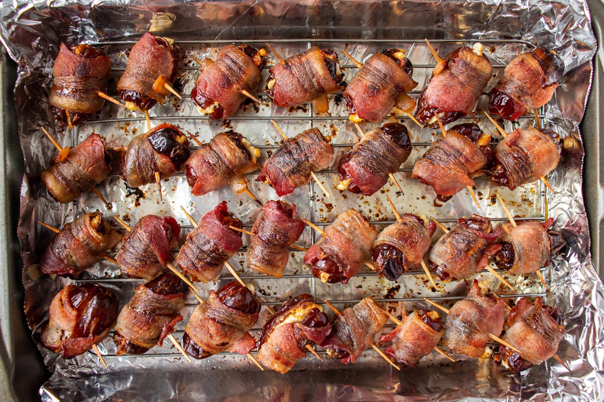 Cooked bacon wrapped dates skewered with toothpicks in a rectangular baking pan.