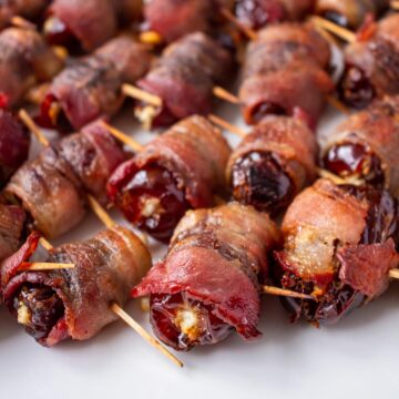 Bacon wrapped dates skewered with toothpicks arranged in rows on a white plate.