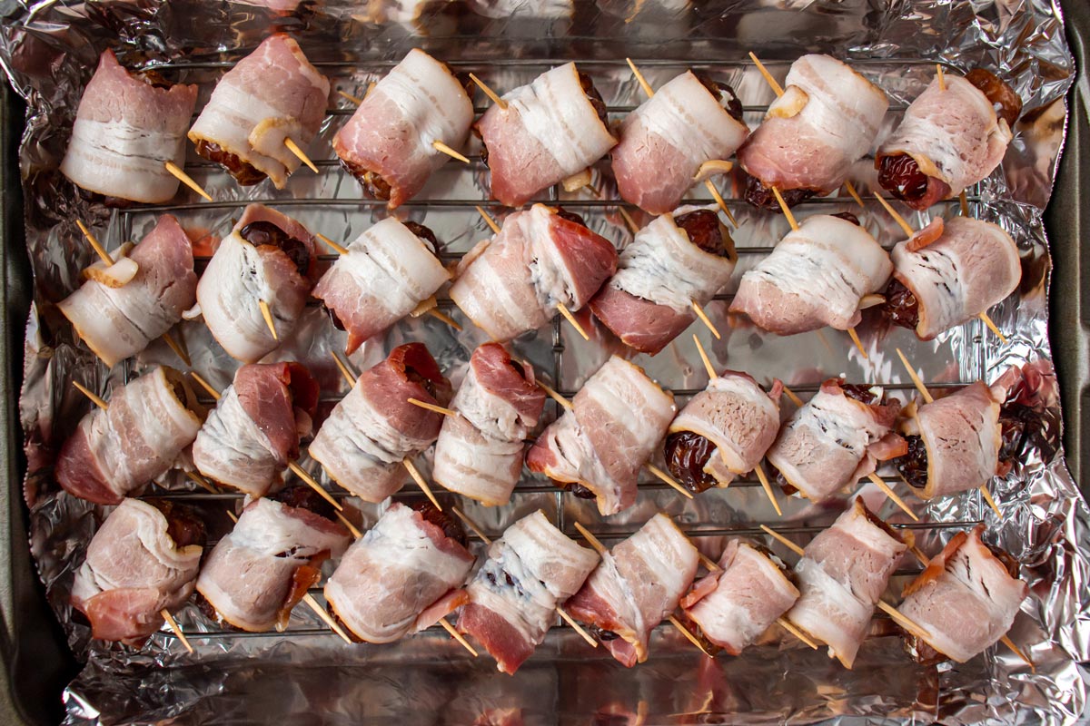 Uncooked bacon wrapped dates skewered with toothpicks in a rectangular baking pan.