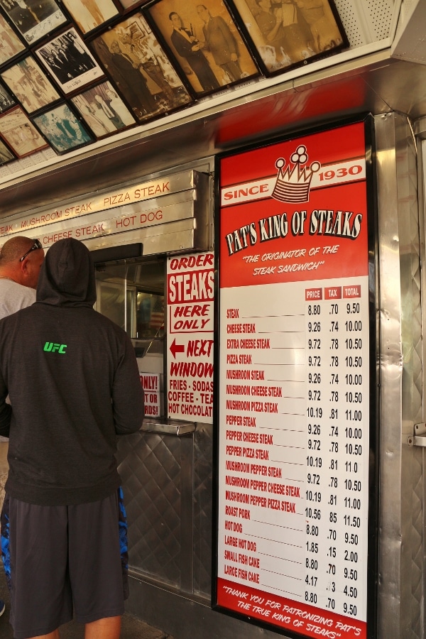 a menu with prices on a wall outside Pat\'s King of Steaks