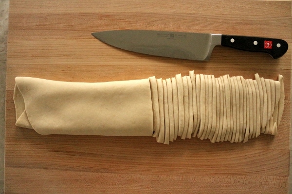 A knife on a wooden cutting board next to dough partially cut into strips