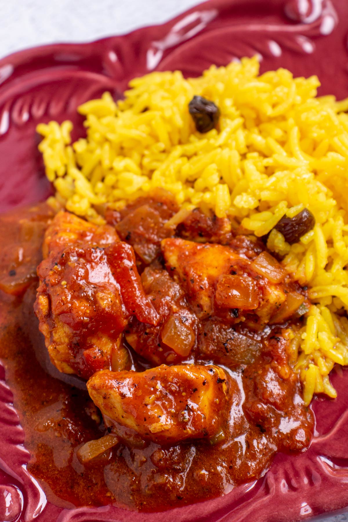 Chicken curry in tomato sauce with yellow rice on a dark red plate.