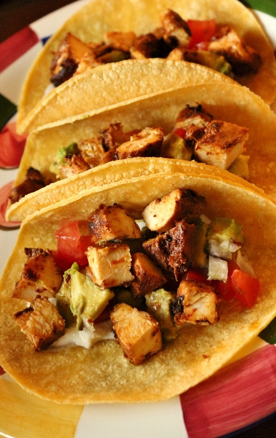 Three soft tacos lined up on a plate with cubed chicken, tomatoes, and guacamole.