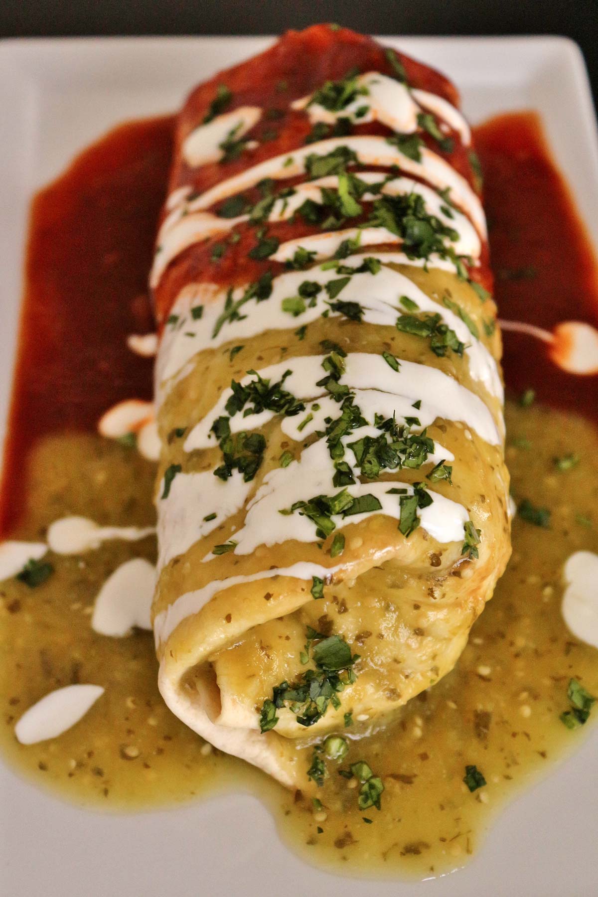 A burrito topped with green, red, and white sauces on a white rectangular plate.