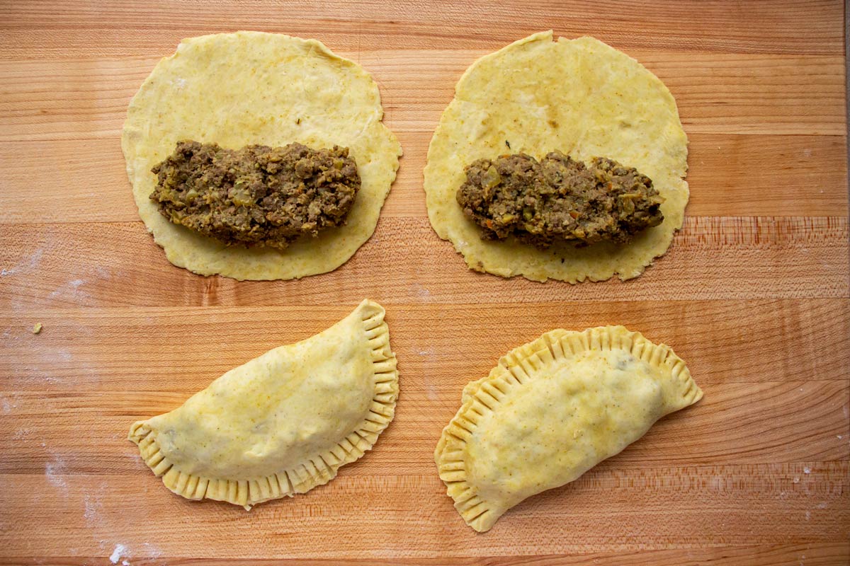 Assembling Jamaican beef patties by folding dough over filling on a wooden board.