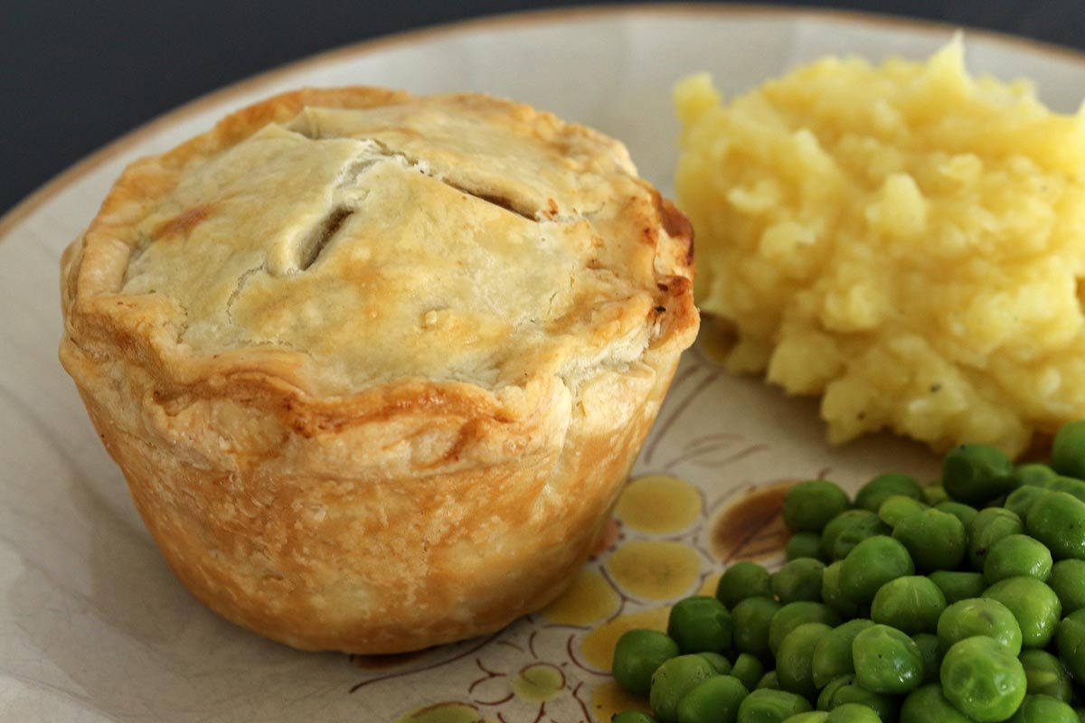 An individual pie on a rustic plate with mashed potatoes and peas on the side.