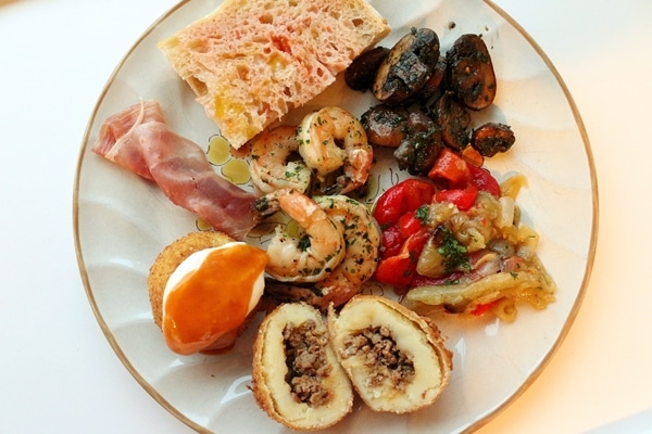 A plate of tapas including pan con tomate, mushrooms, shrimp, roasted vegetables, ham, and potato balls