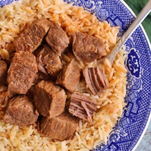 Cubes of stewed beef on top of rice pilaf in a shallow blue and white bowl.
