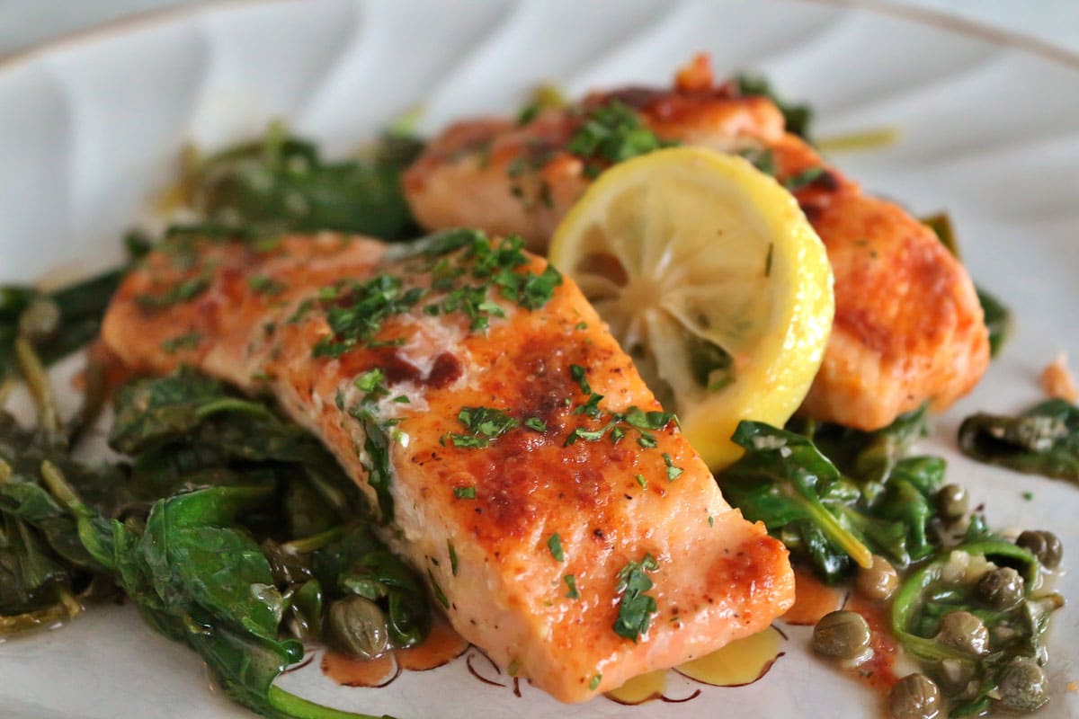 A plate of cooked salmon fillets with wilted spinach, capers and lemon slices.