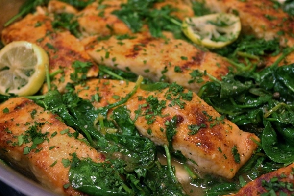 Salmon piccata fillets cooking in a pan with wilted baby spinach and lemon slices