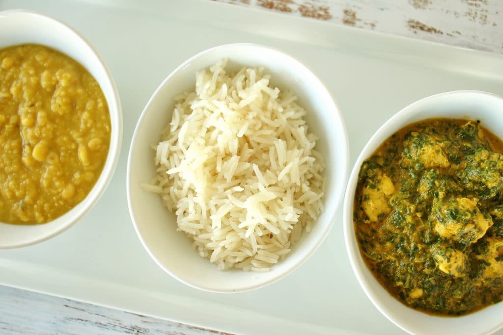 A row of 3 small white bowls. From left to right, they are filled with chana dal (a golden colored Indian lentil dish), basmati rice, and chicken saag (spinach curry).