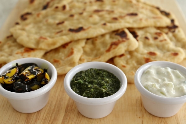 Three Sanaa dipping sauces in little cups on a wooden board with a pile of naan in the background
