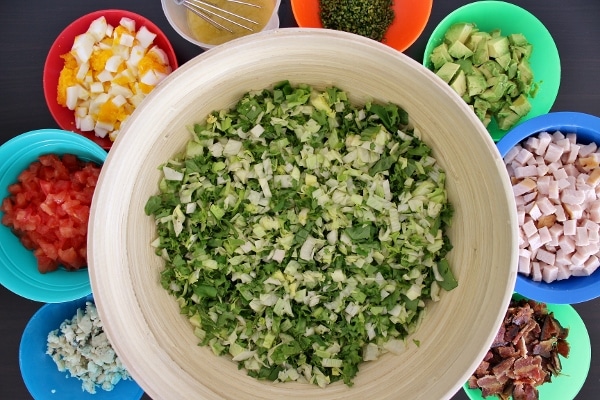 Chopped greens in a large salad bowl surrounded by smaller bowls filled with toppings for Cobb salad: crumbled blue cheese, chopped tomatoes, hard-boiled eggs, dressing, chives, avocado, chopped turkey, and bacon.