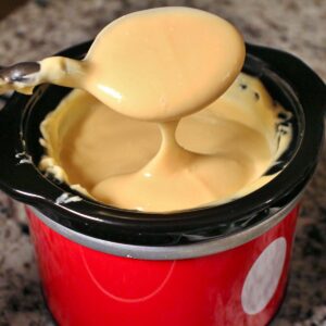 Homemade nacho cheese sauce in a small red warmer with a spoon lifting some out.