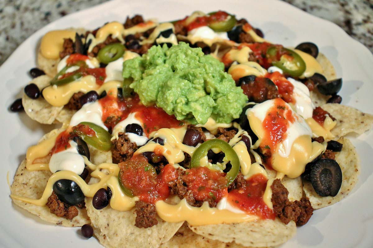 A plate of loaded nachos with cheese sauce, black beans, beef, olives, sour cream, and guacamole.