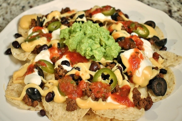A plate of loaded nachos with homemade nacho cheese sauce, taco beef, pickled jalapenos, black olives, black beans, guacamole, salsa, and sour cream