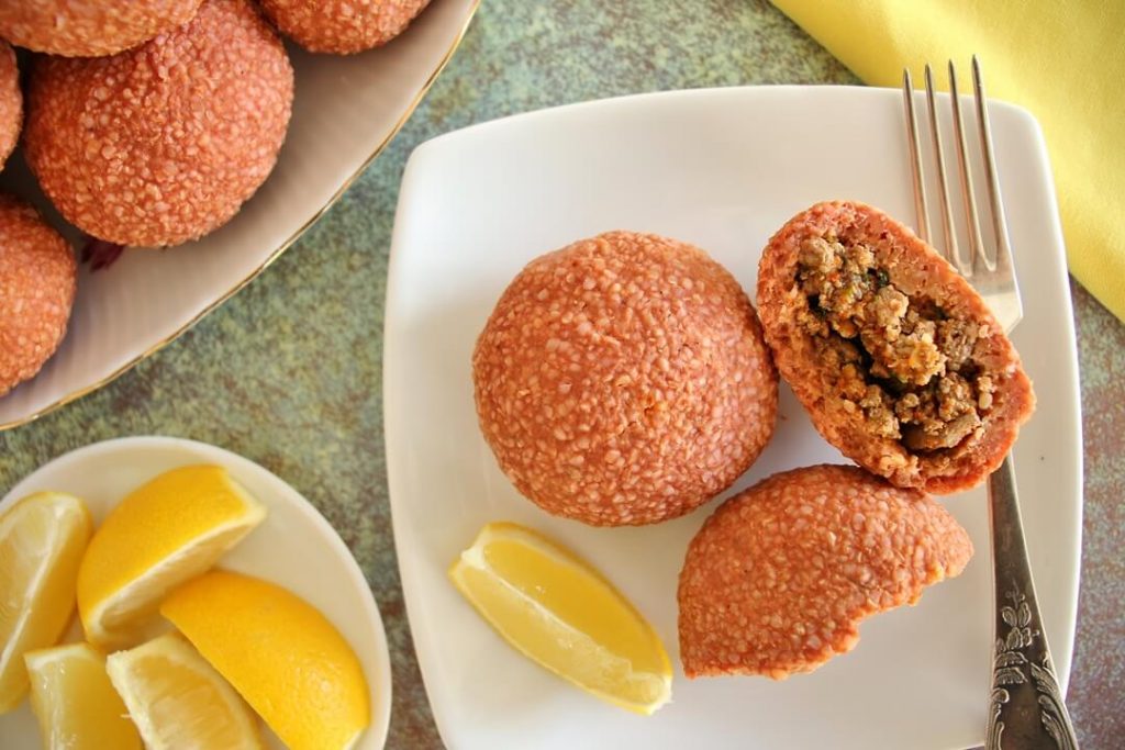 2 round ishli koftes on a white square plate, 1 is broken in half to show the filling. There is a lemon wedge on the plate, and a plate of lemon wedges and platter of more koftes next to the plate.