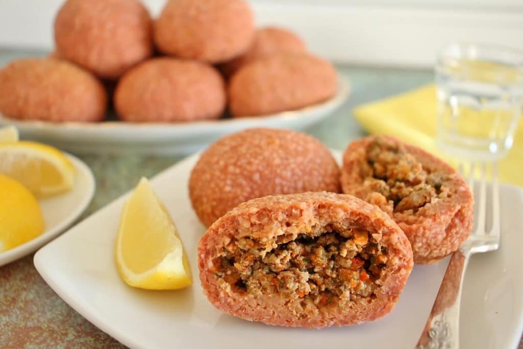 2 round ishli koftes on a white square plate, 1 is broken in half to show the filling. There is a lemon wedge on the plate, and a plate of lemon wedges and platter of more koftes next to the plate.