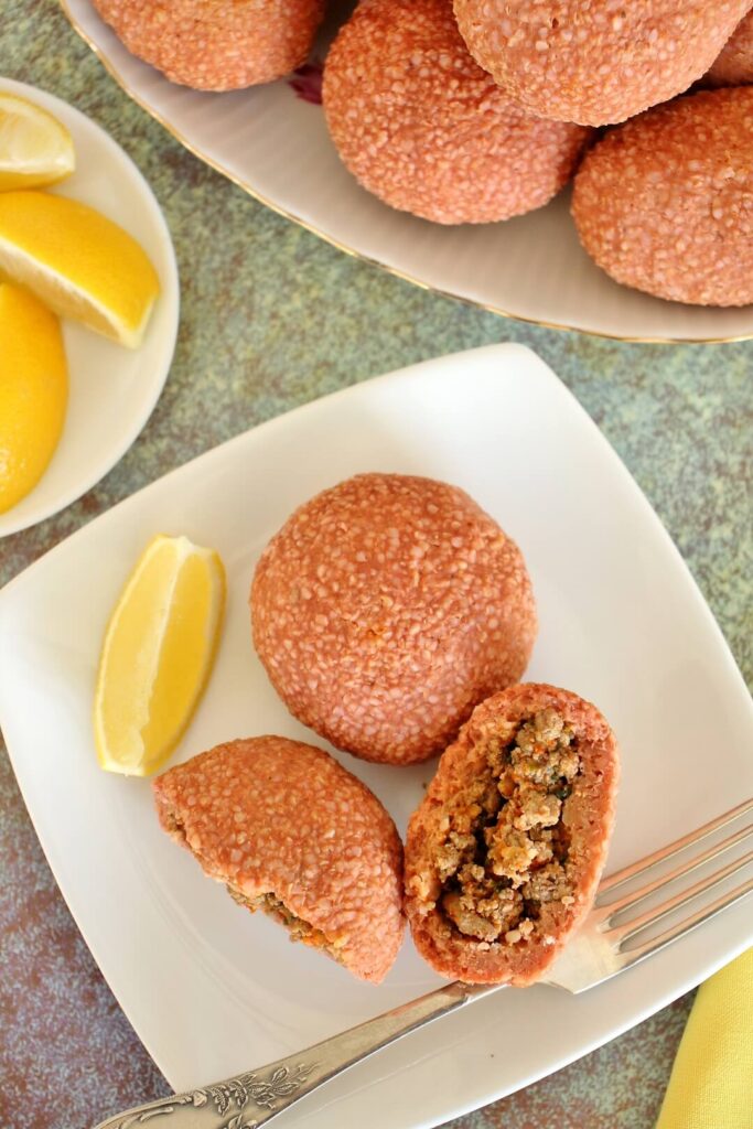 2 round ishli koftes on a white square plate, 1 is broken in half to show the filling. There is a lemon wedge on the plate, and a plate of lemon wedges and platter of more koftes next to the plate.