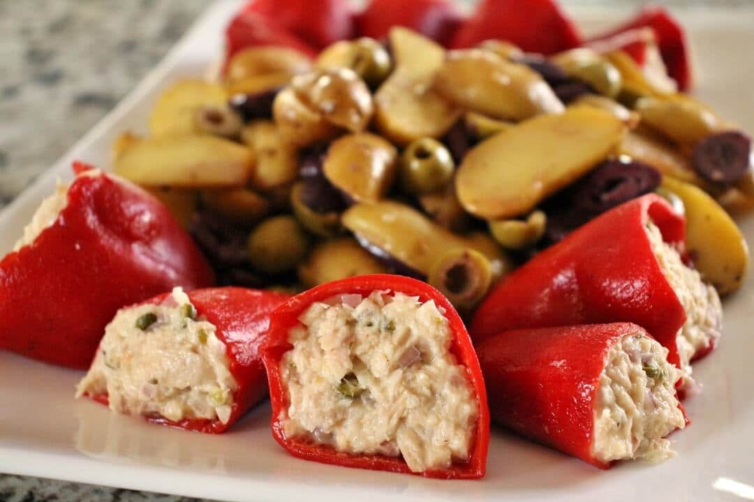 Five piquillo peppers stuffed with tuna salad, with a large serving of potato and olive salad behind it on the plate.