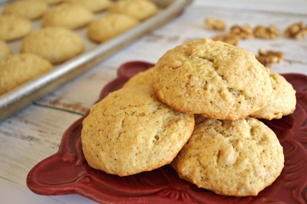 Five cakey yogurt cookies on a square dark red plate, with a tray of cookies in the background