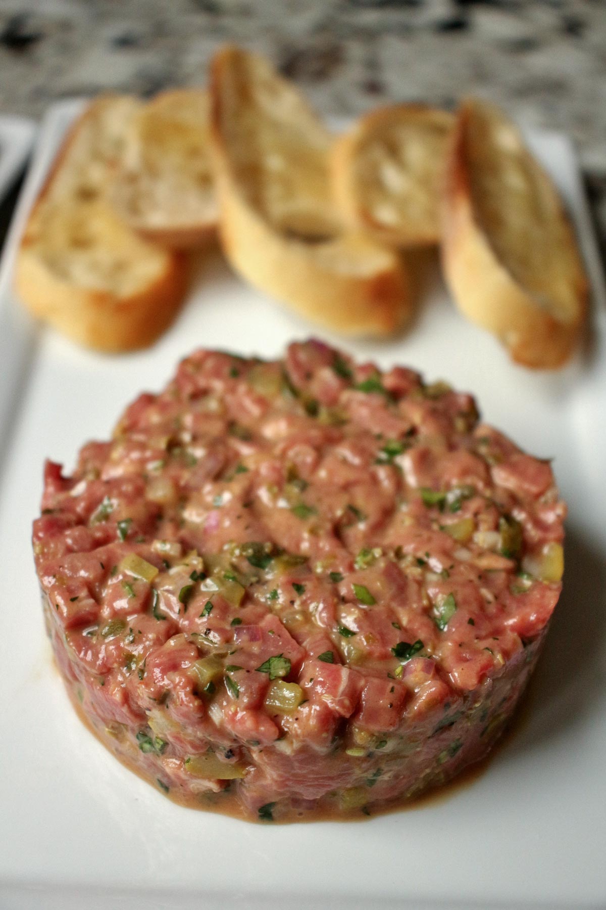 Closeup of steak tartare in round, flat patty, served on a while rectangular plate.