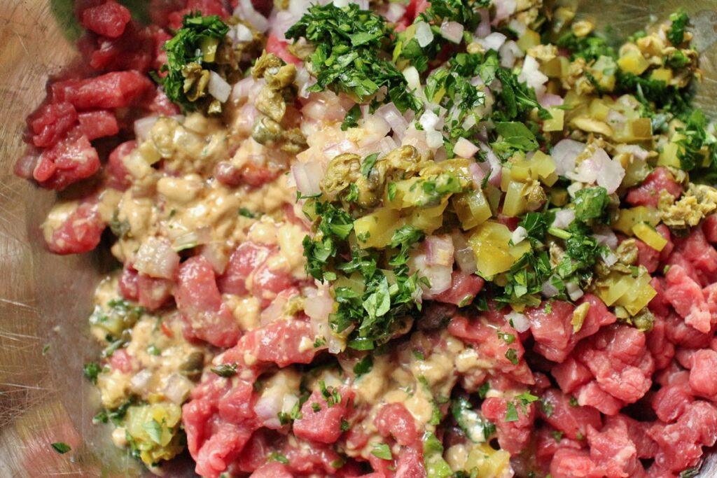 Chopped beef tenderloin in a mixing bowl topped with chopped pickles, parsley, capers, shallots, and a Dijon dressing