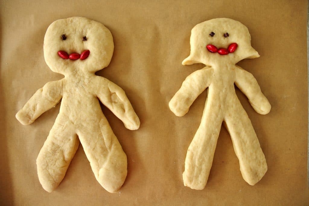 A man and woman chorek person on a parchment lined baking sheet, with whole cloves for eyes and red m&ms for mouths, prior to baking