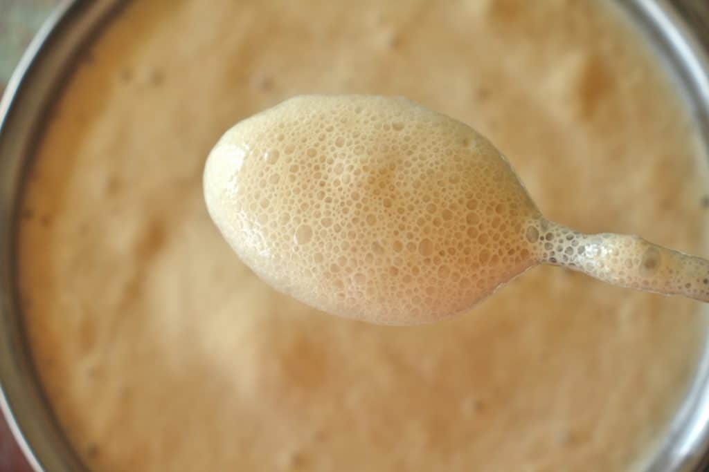 Frothy yeast bloomed in water with sugar, with a spoon holding some up to show the bubbles