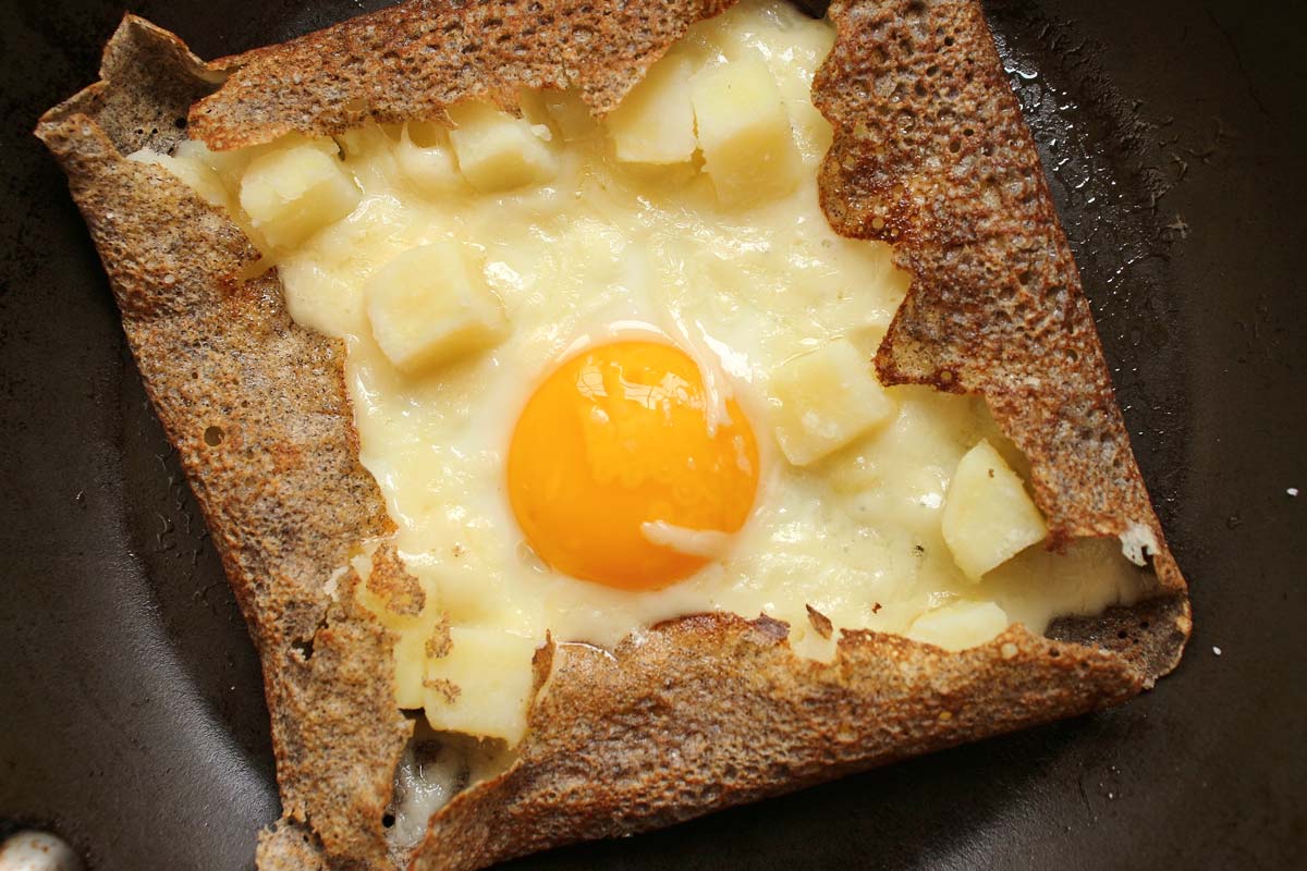 A buckwheat galette filled with potatoes, cheese, and fried egg in a frying pan