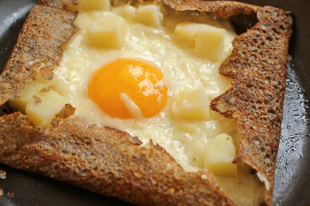 A buckwheat galette filled with potatoes, cheese, and fried egg in a frying pan