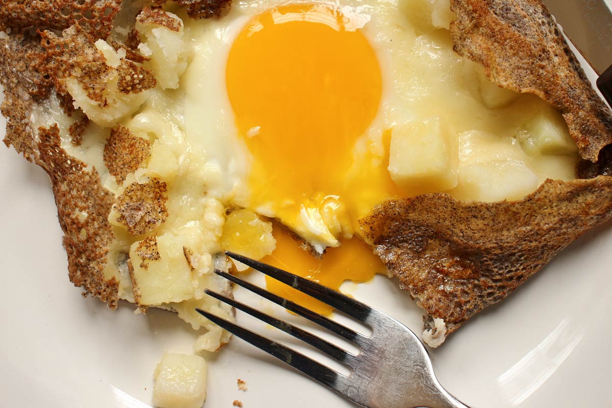 A buckwheat galette filled with potatoes, cheese, and fried egg, yolk oozing onto the plate