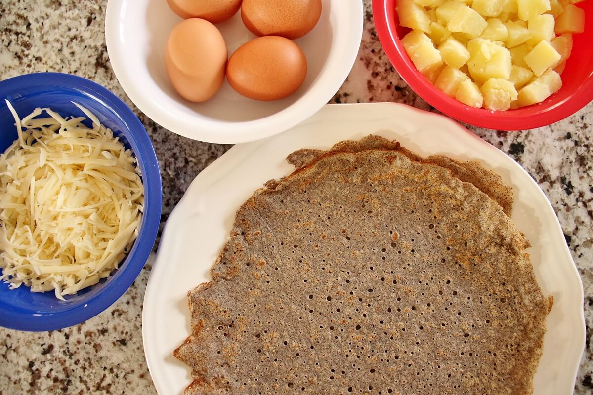 stack of buckwheat galettes, bowls of shredded cheese, uncracked eggs, cooked cubed potato