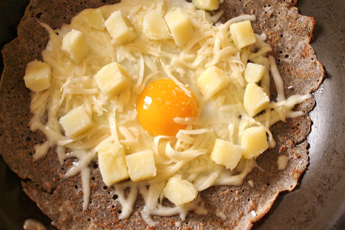 buckwheat crepe in a frying pan topped with an egg, shredded cheese, and cubed potato