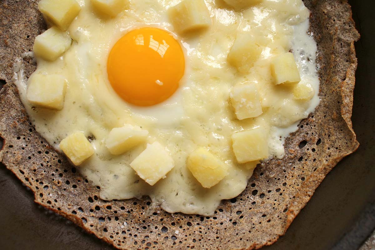 buckwheat crepe in a frying pan topped with a cooked egg, melted cheese, and cooked cubed potatoes
