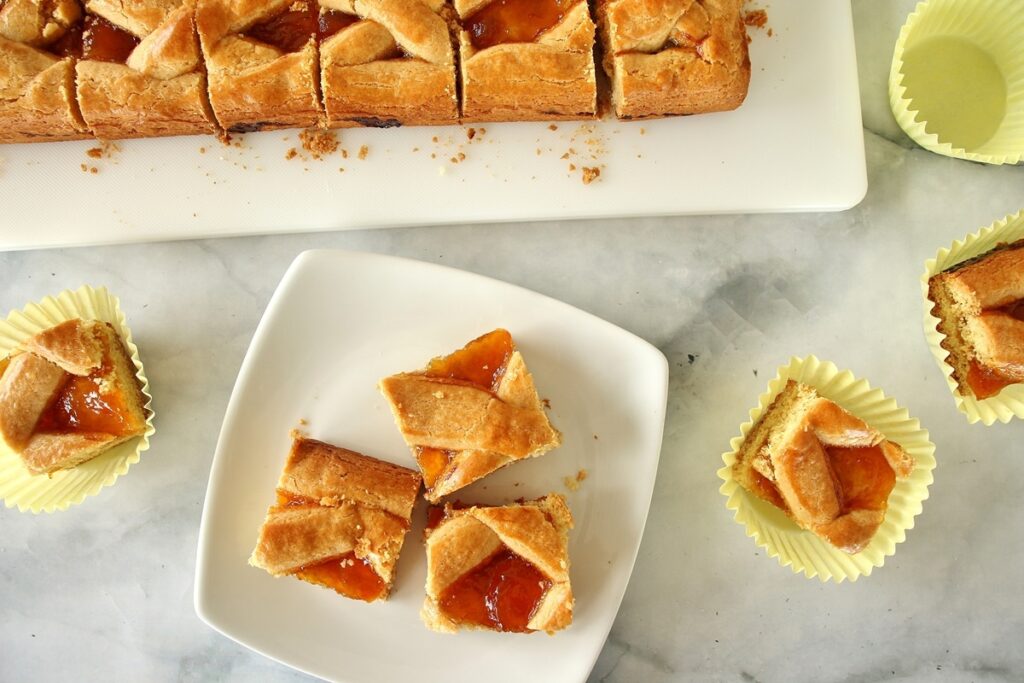 Russian pirog cut into squares on a cutting board to the top of the frame. Three squares of lattice-topped pirog are on a white square plate, with 3 other squares in muffin liners on the marble countertop.