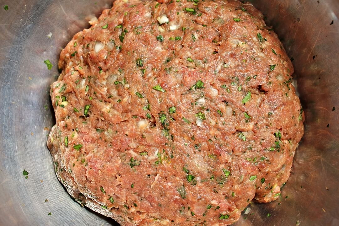 Ground beef mixed with chopped onions, parsley, and seasonings in a metal mixing bowl