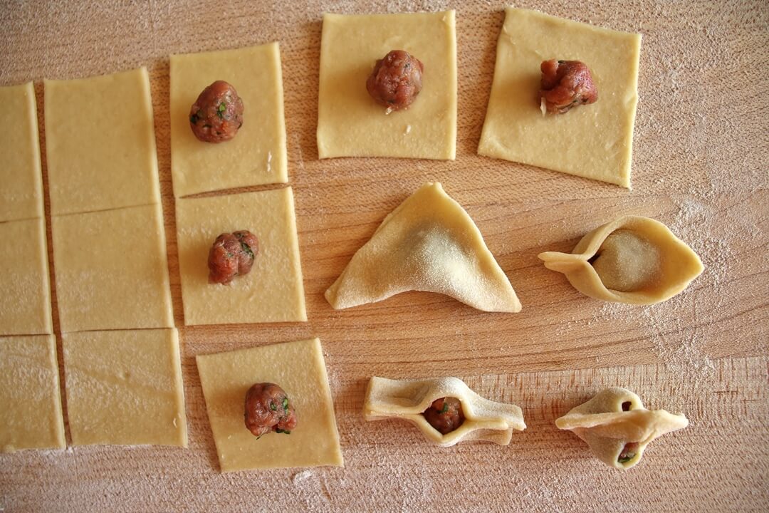 a wooden board covered with squares of dough being shaped into manti dumplings