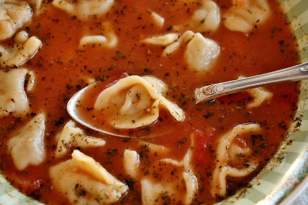 a bowl of manti soup with a spoon lifting a dumpling out of the tomato broth