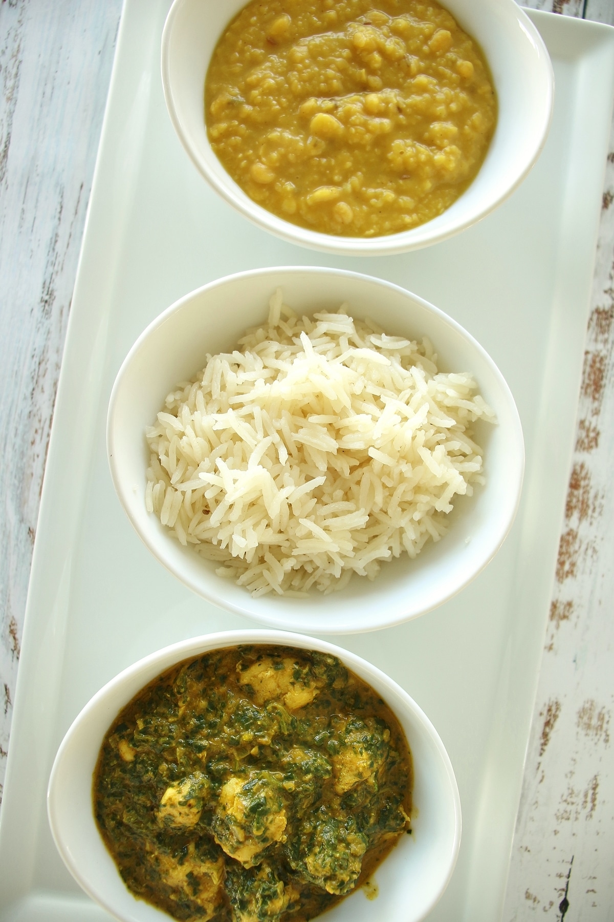 A row of 3 small white bowls filled with yellow and green curries and rice.