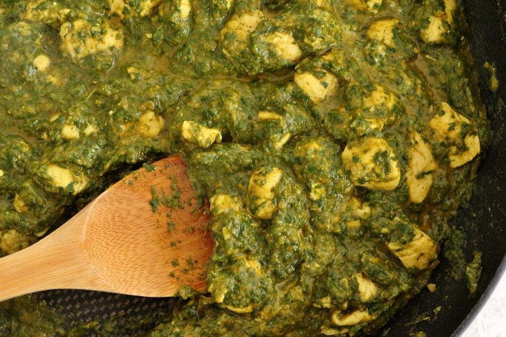 Cooking chicken saag in a frying pan, with a wooden spoon stirring the mixture