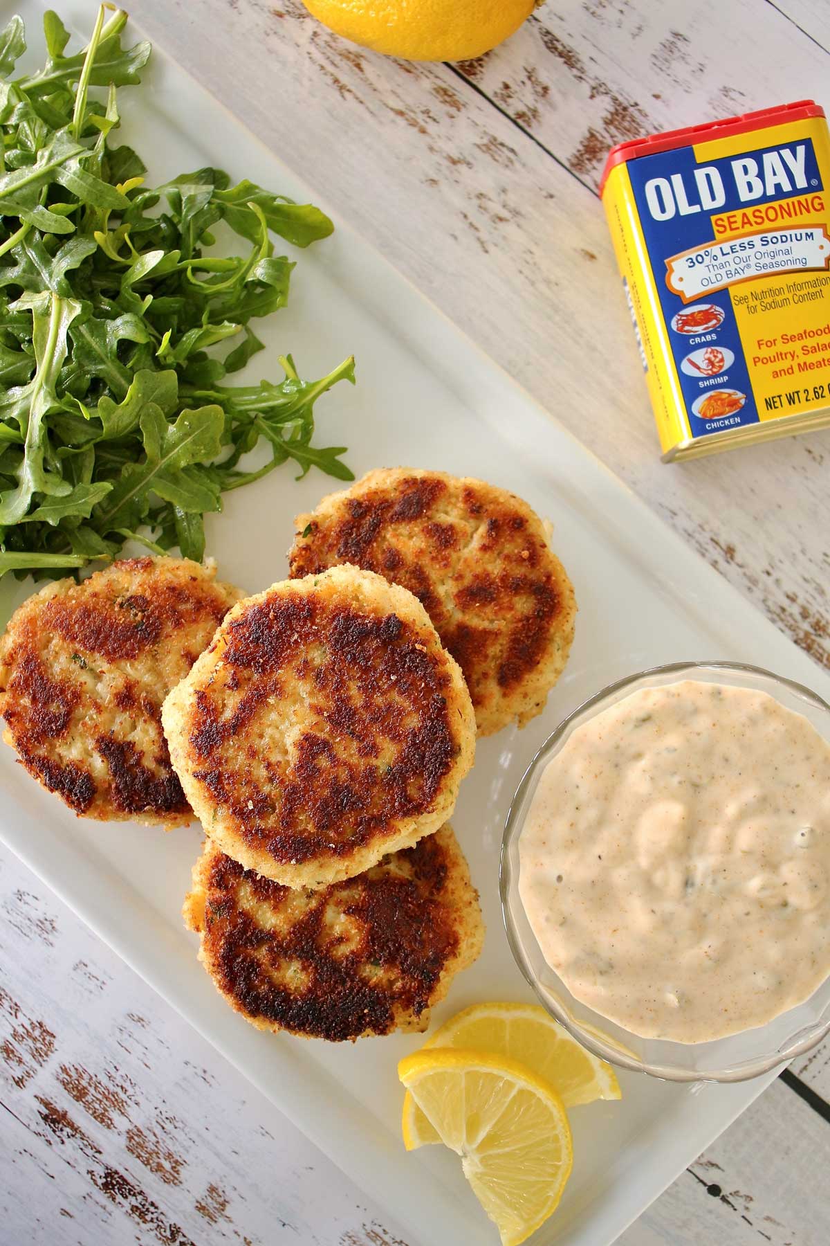 4 crab cakes on white plate, a bowl of sauce, and arugula salad