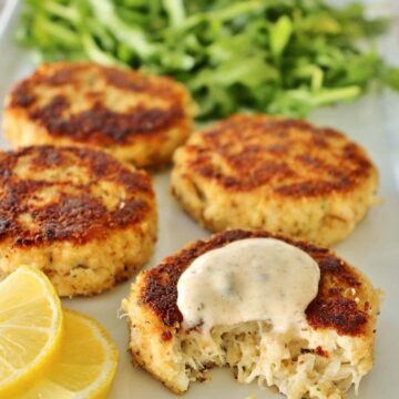4 crab cakes with lemon slices and arugula on white plate