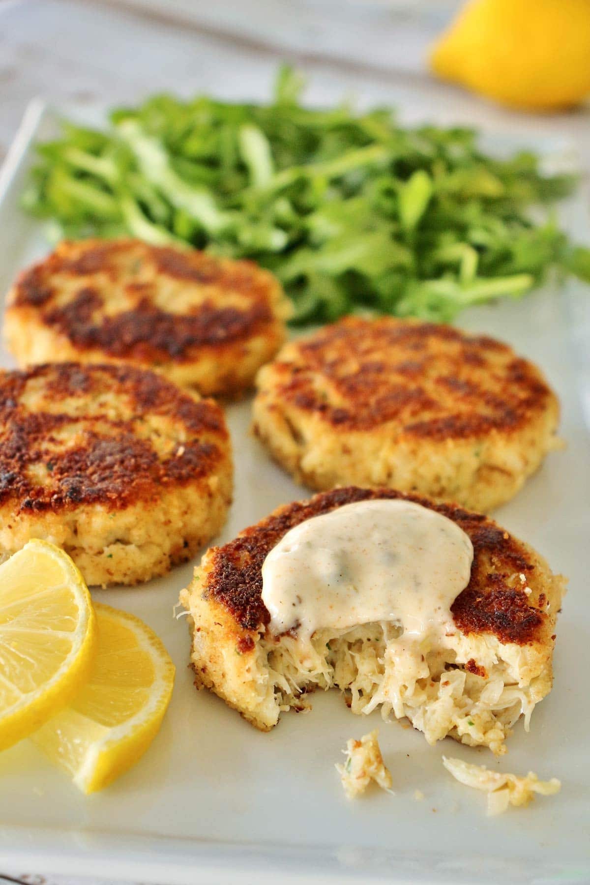 4 crab cakes with lemon slices and arugula on white plate