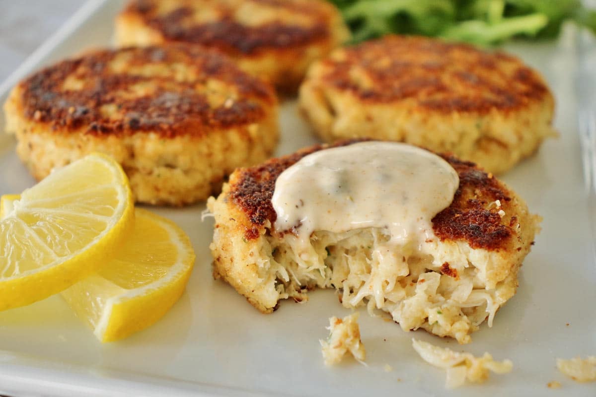 4 crab cakes on white plate with dollop of sauce on 1 with bite taken out