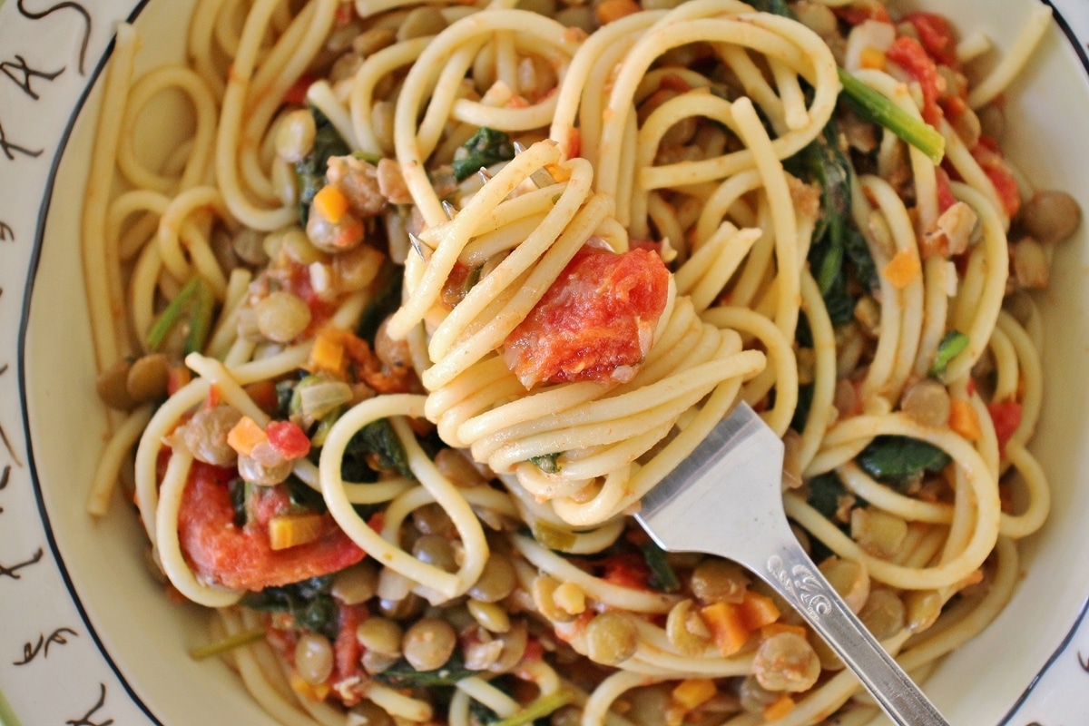 A bowl of spaghetti with lentils, roasted tomatoes, and spinach, with a fork twirled around a portion of pasta