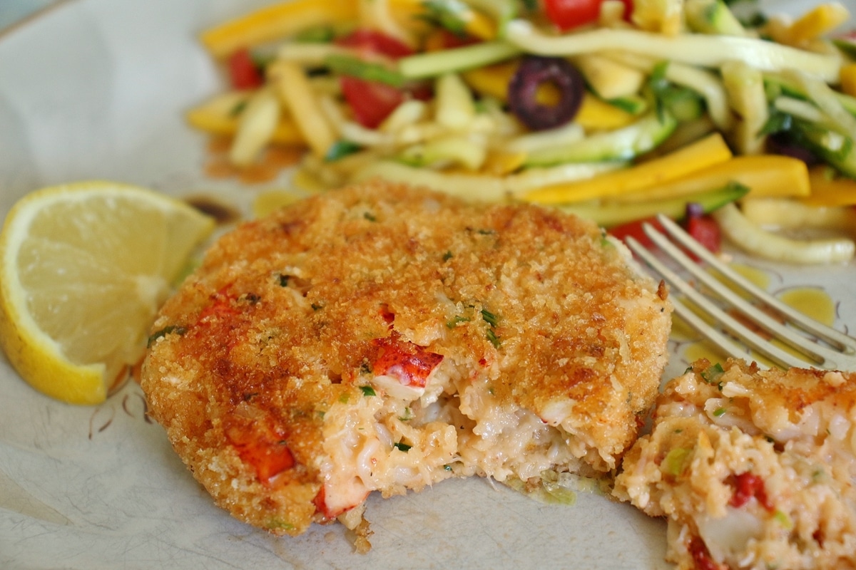 A lobster cake with a bite taken out, served with a lemon wedge and zucchini slaw