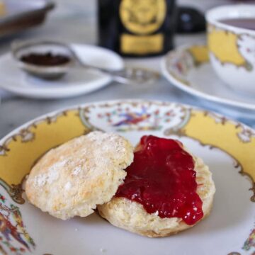 a tea scone topped with jam, served on china with a cup in the background